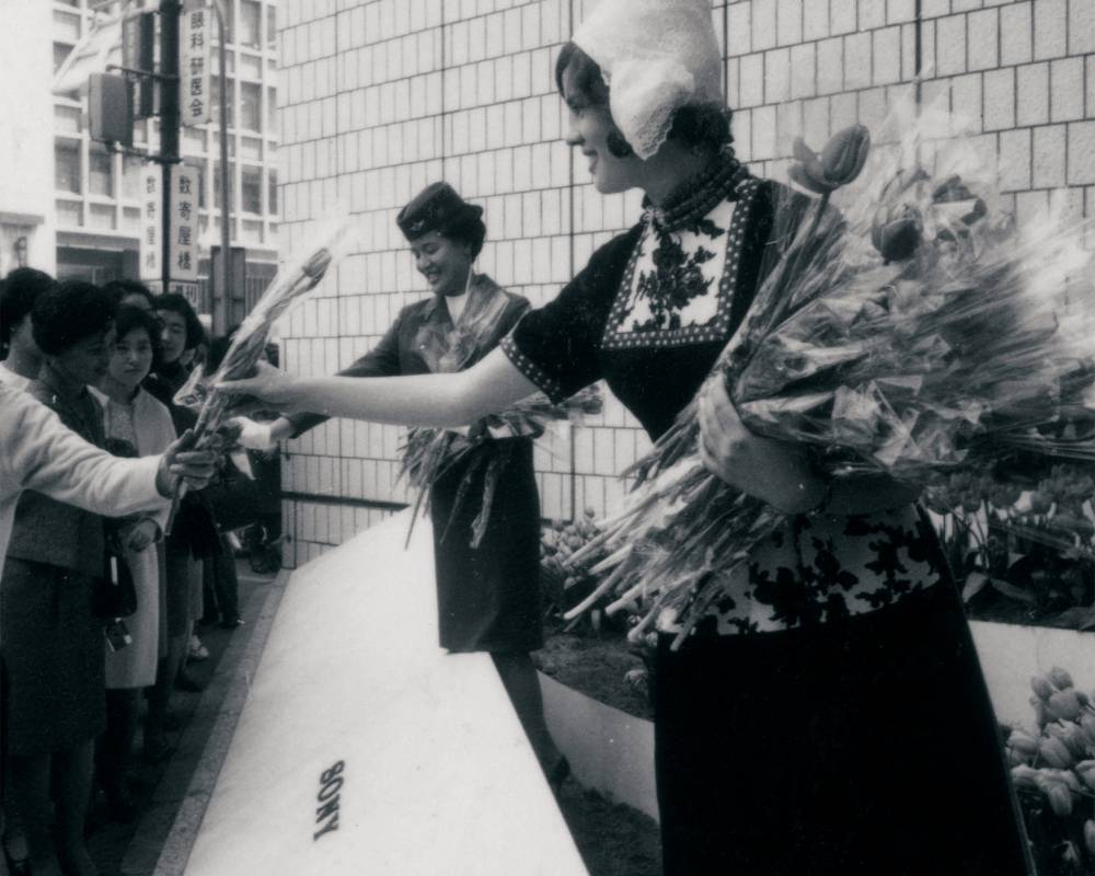 イベントの様子（モノトーン）。ソニースクエア（屋外イベントスペース）でオランダの民族衣装を着た女性と客室乗務員がチューリップを配布している。