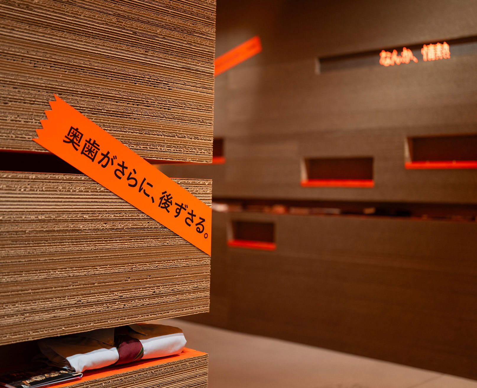 展示風景。地層のような壁面に、「奥歯がさらに、後ずさる。」と書かれたオレンジ色のテープが貼られている。