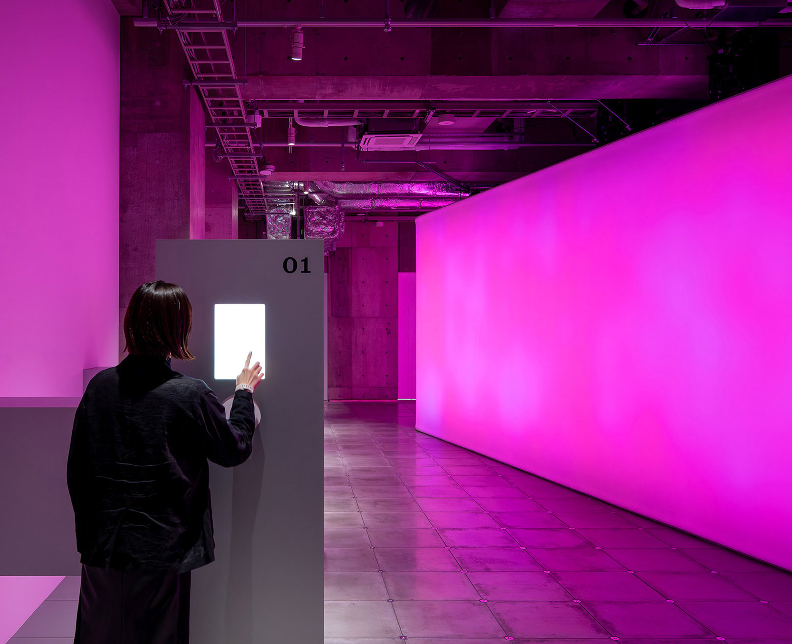 展示風景。ピンク色にライトアップされた展示フロアで、インタラクティブなディスプレイに触れる人。