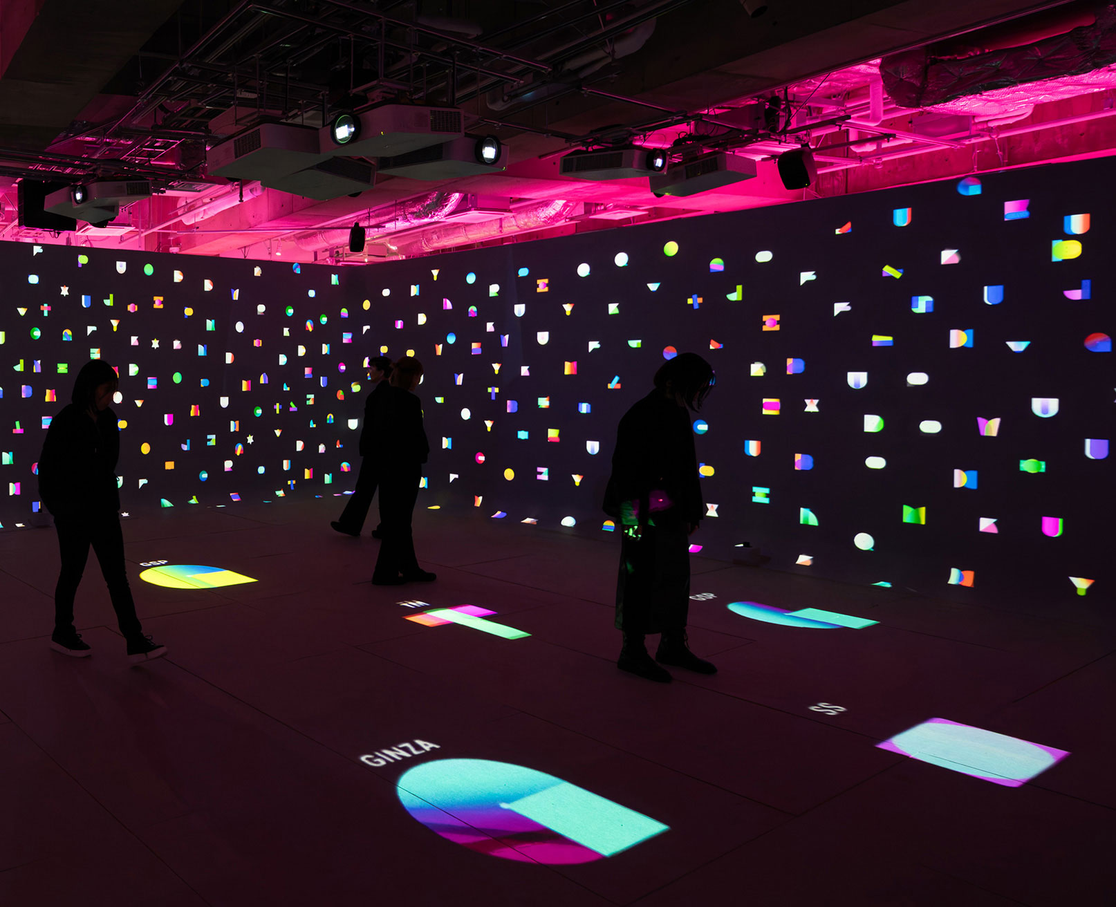 展示風景。暗い空間で、色とりどりのオブジェクトが壁や床に映し出され、人々がその中を歩いている。