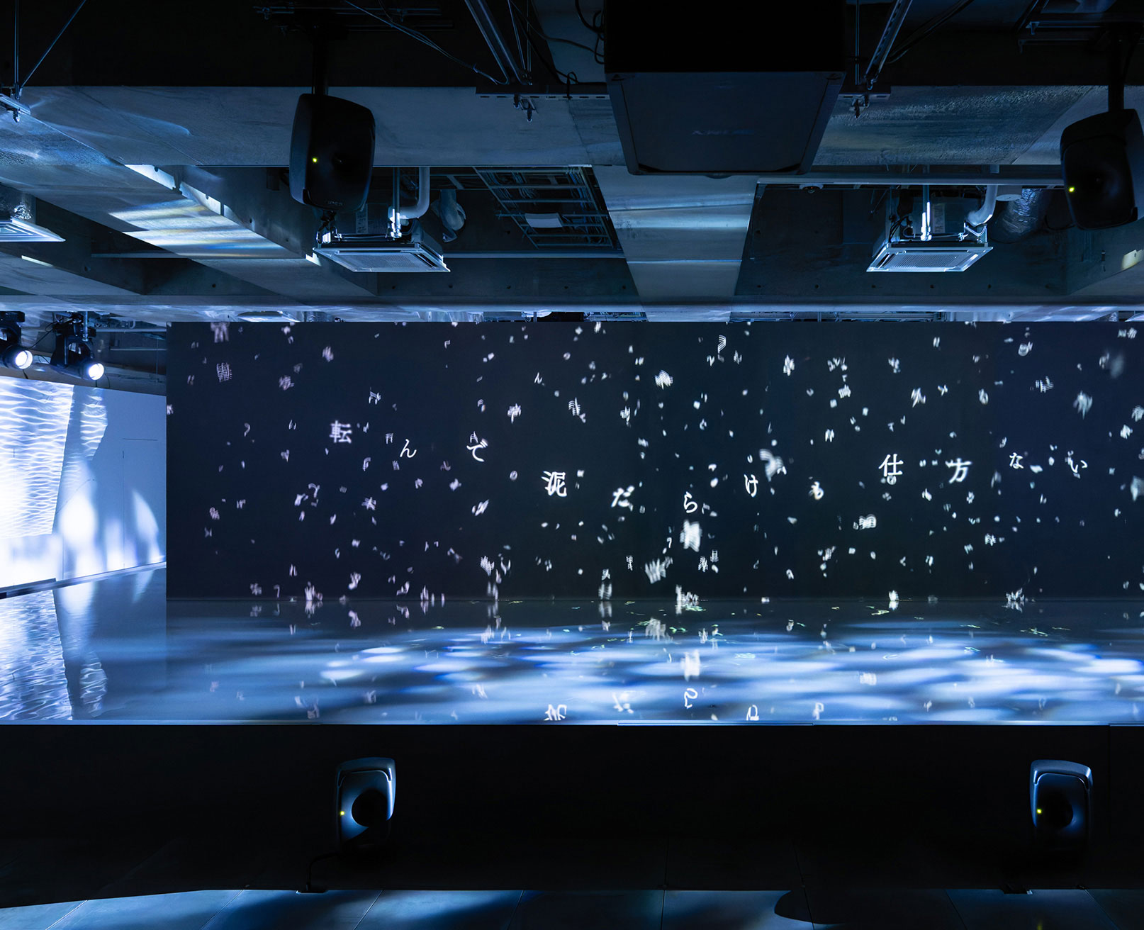 展示風景。黒い壁面にプロジェクションされるさまざまな言葉と、その手前のある水面と光の反射によるインスタレーション。