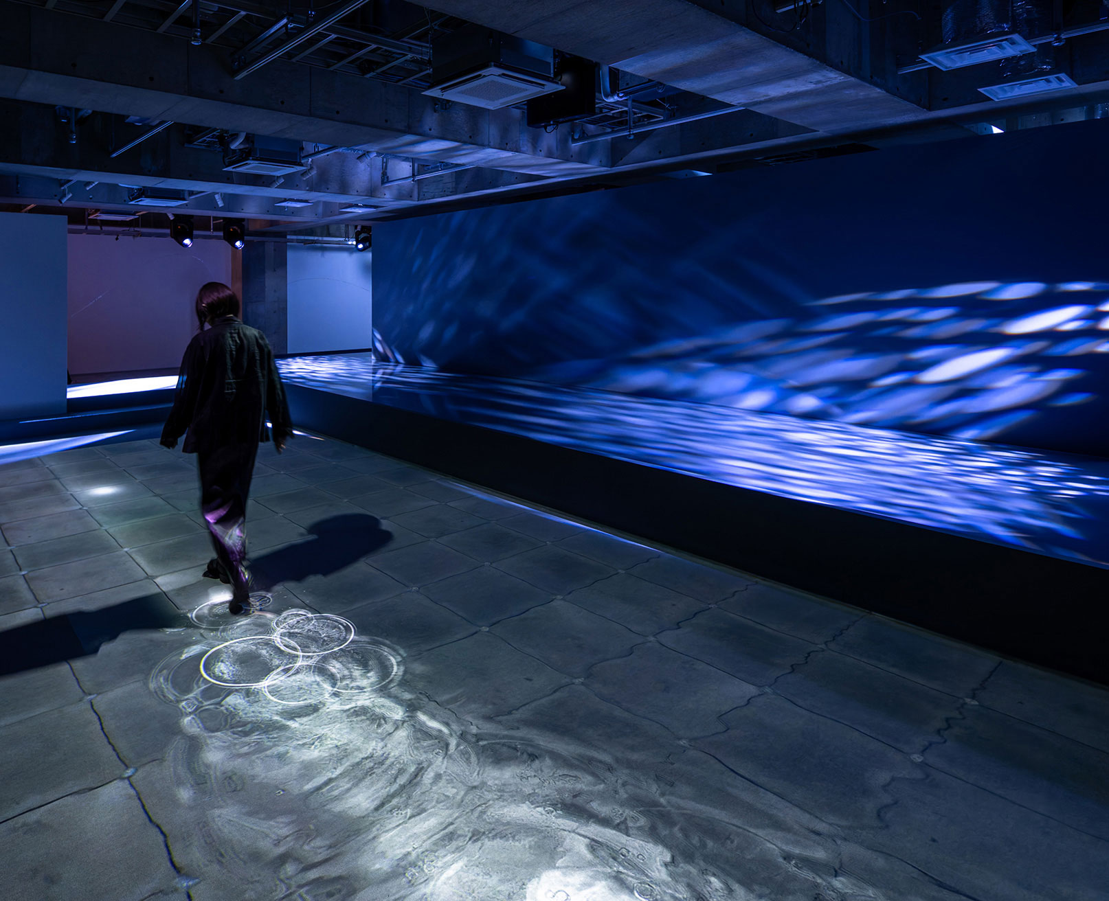 展示風景。青い光に包まれた空間で、波紋のパターンが床に投影されており、その上を歩いている人の姿が見える。