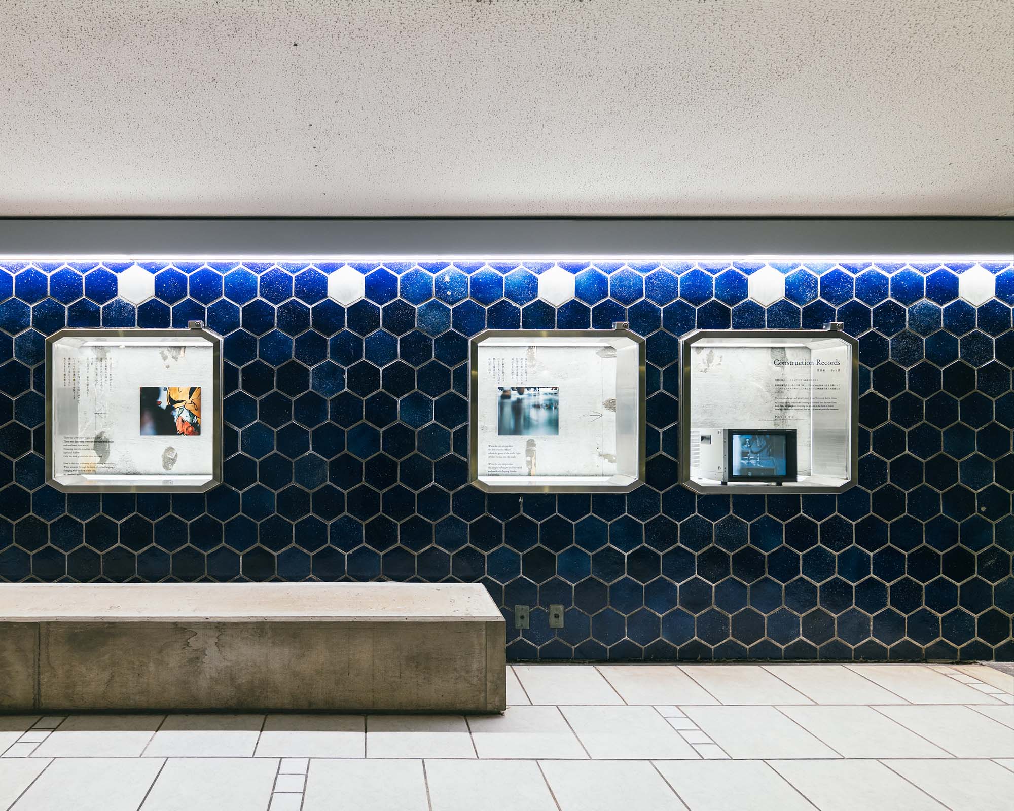 Display cases set into a hexagonal-tiled wall on the B1 floor of Ginza Sony Park, presenting photos and texts that reveal fragments of memory and landscape.