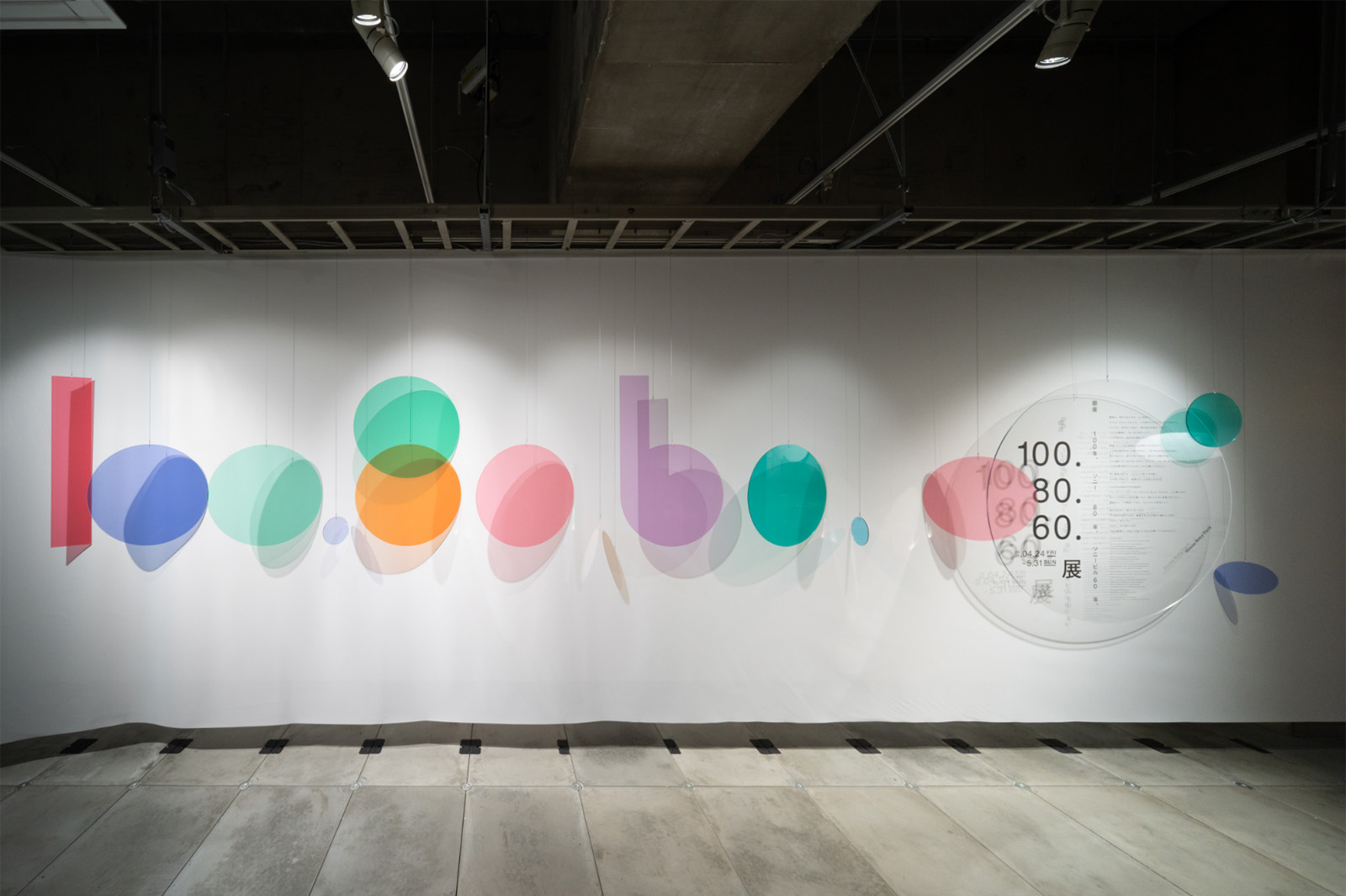 Introductory visual of the exhibition. Colorful circular graphics line the wall alongside the “100. 80. 60. Exhibition.” logo, presenting the concept of the exhibition.