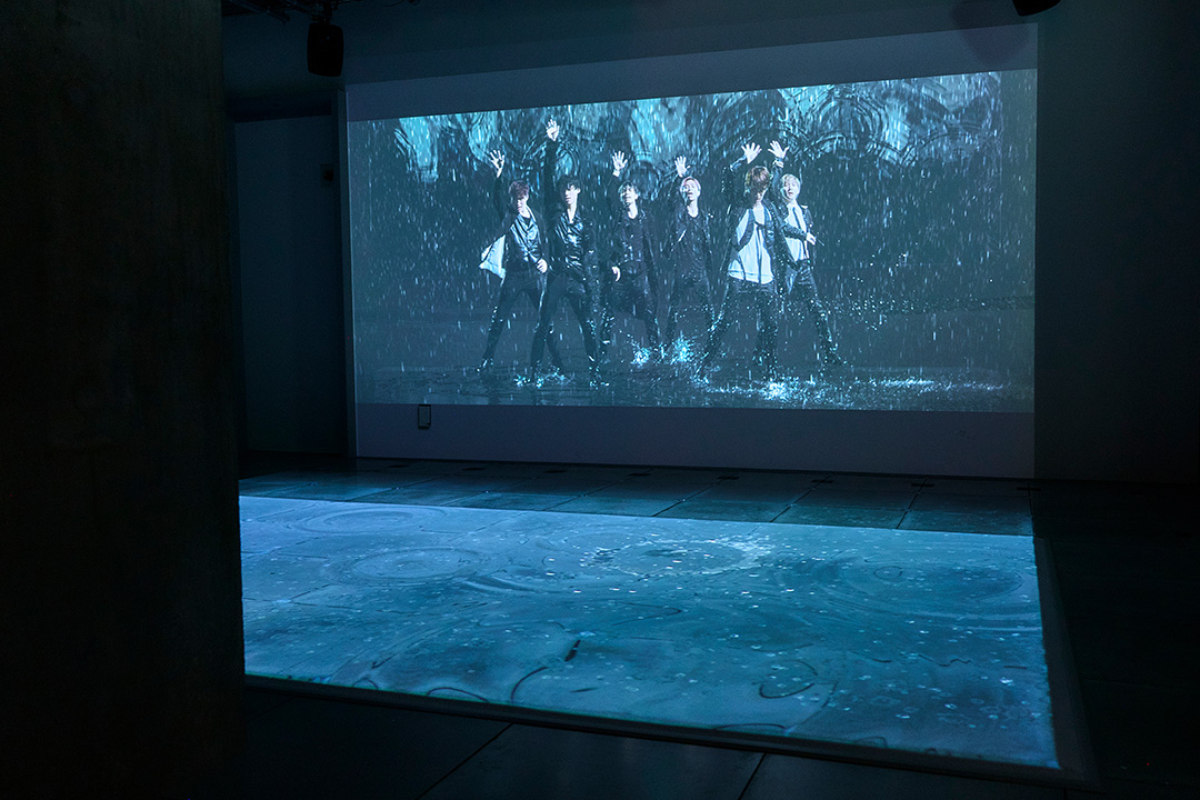 薄暗い展示室の大型スクリーンに、激しい雨の中でパフォーマンスを行うSixTONESの映像が投映されている。スクリーンの手前にある床面にも、雨粒や波紋の映像が映し出されており、部屋全体が幻想的な水の世界に包まれている。