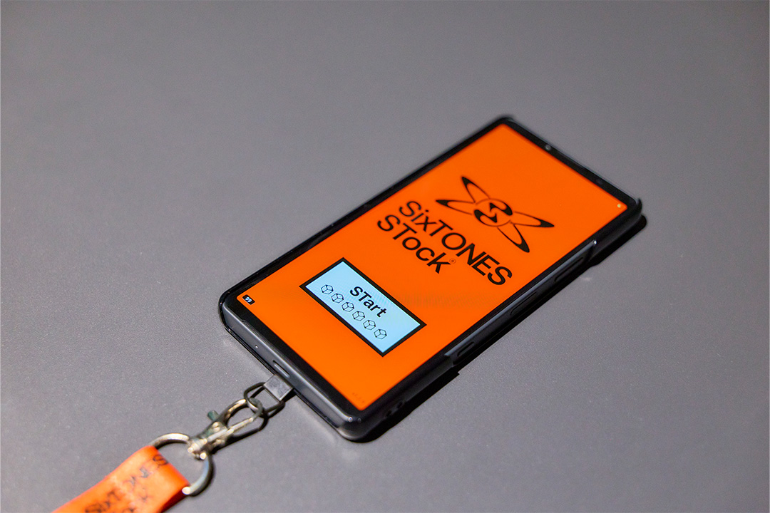 グレーの台座の上に斜めに置かれたスマートフォン。鮮やかなオレンジ色の画面には、黒い「SixTONES STock」のロゴマークと「STart」と書かれた青いボタンが表示されている。スマホの下部には、オレンジ色のネックストラップが金具で取り付けられている。