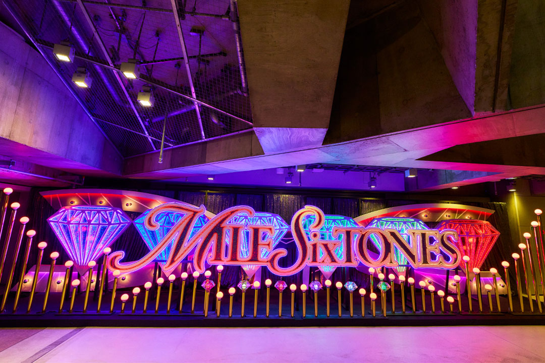 薄暗くスタイリッシュなコンクリート空間に飾られた、「MILESixTONES」のネオンロゴオブジェ。ネオンの文字の背景にはカラフルに輝く大きなダイヤモンド型のライトが配置され、手前には小さな球体照明がずらりと一列に並んで輝いている。