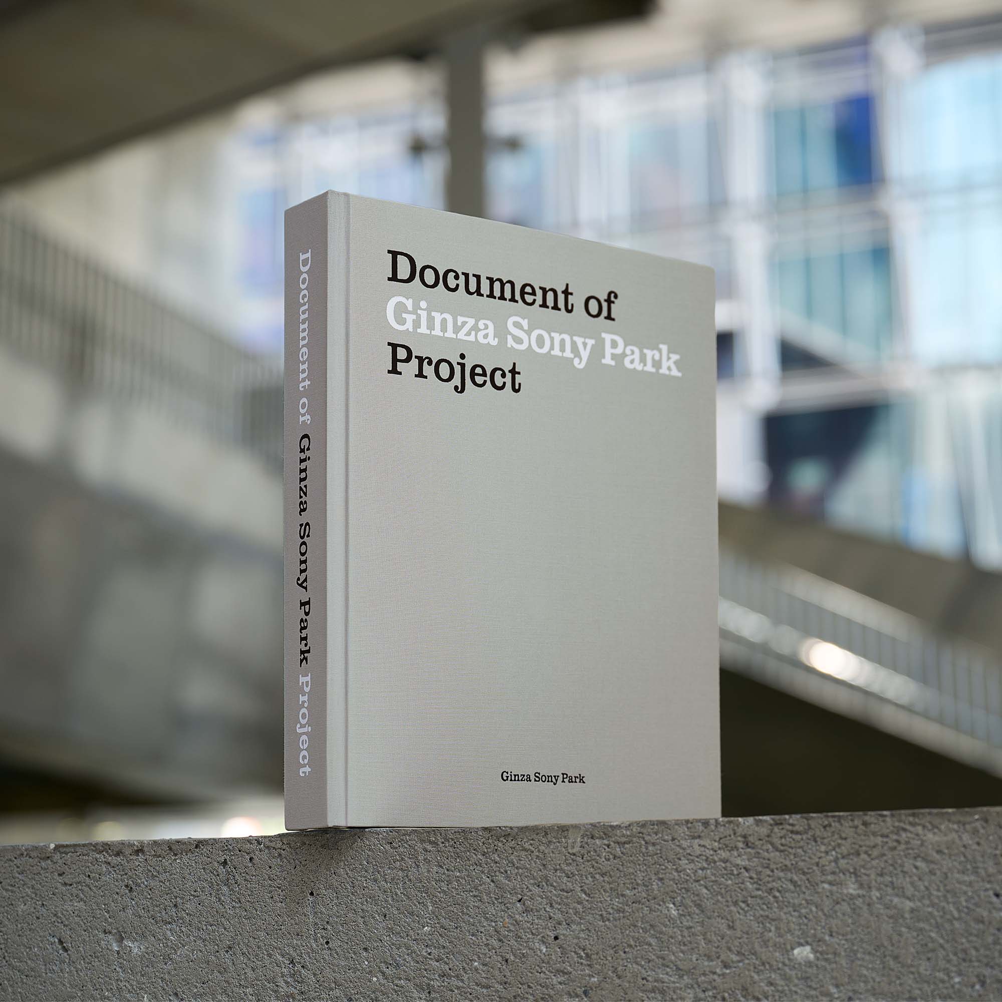 コンクリートの構造物の上に自立するグレーの書籍。表紙と背表紙に「Document of Ginza Sony Park Project」と書かれており、背景にはぼやけたビルの屋内空間が写っている。