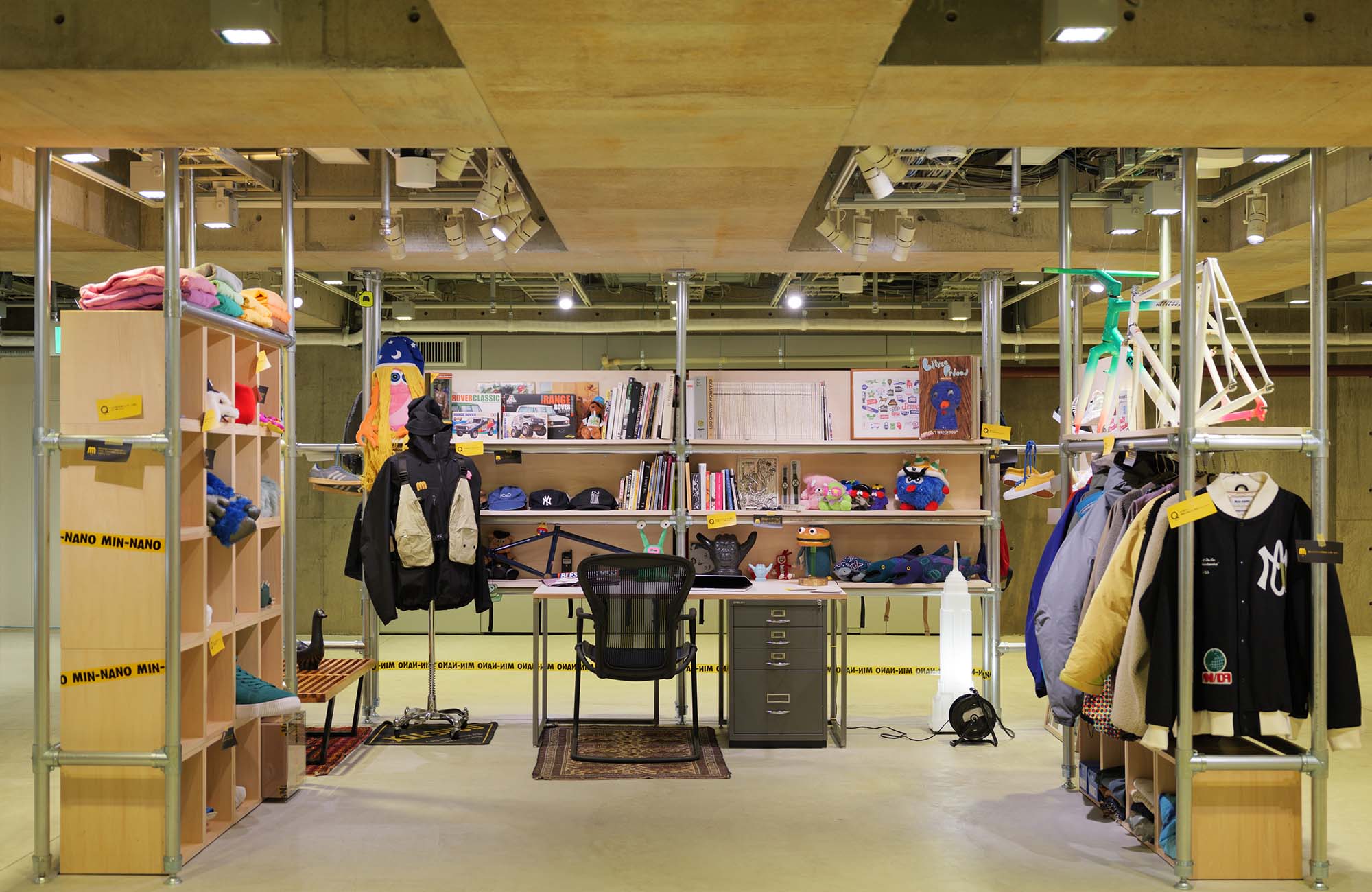 Ginza Sony Park B2の『SHELF PORTRAIT #01 MIN-NANO』より。金属パイプや木製棚で組まれたラックにアウターやキャップ、ぬいぐるみ、書籍、自転車フレームが並ぶ。中央にはデスクとチェアが置かれている。