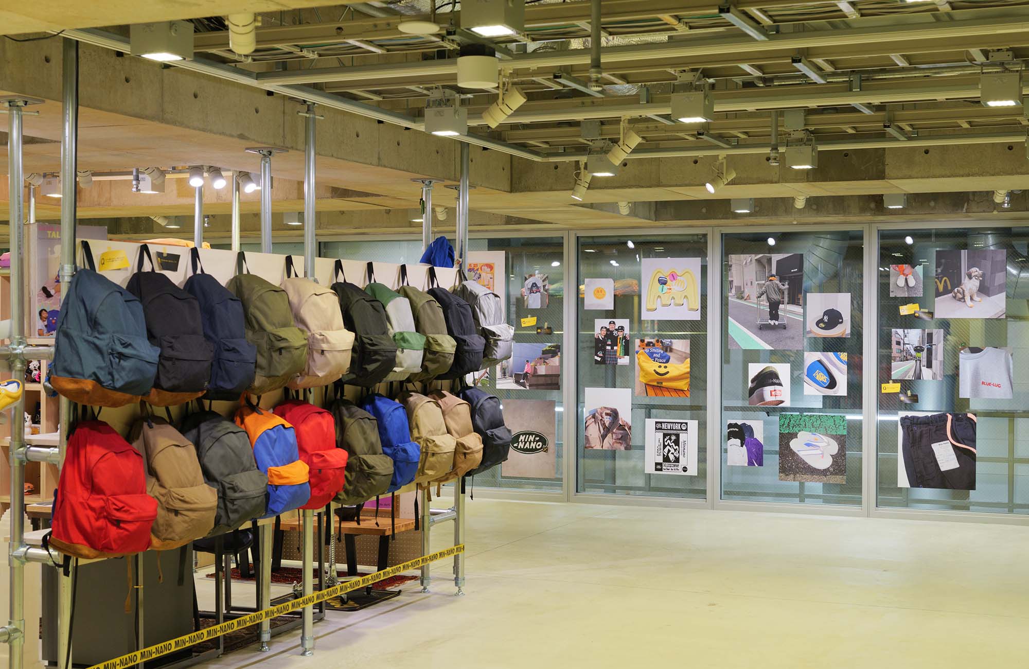Ginza Sony Park B2で開催中の『SHELF PORTRAIT #01 MIN-NANO』の展示風景。金属パイプ用ラックには、カラフルなバックパックが2段に渡って多数吊り下げられている。奥のガラス壁面には、写真が整然と並んで貼られている。