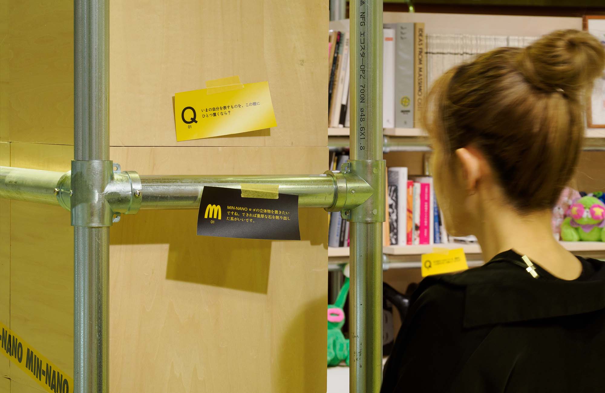 『SHELF PORTRAIT #01 MIN-NANO』の展示棚の側面。質問が書かれた黄色いカード「Q」と、回答が記された黒いロゴ入りカードがパイプフレームを挟んで貼られており、来場者がその解説カードを近くでじっくりと読み込んでいる。