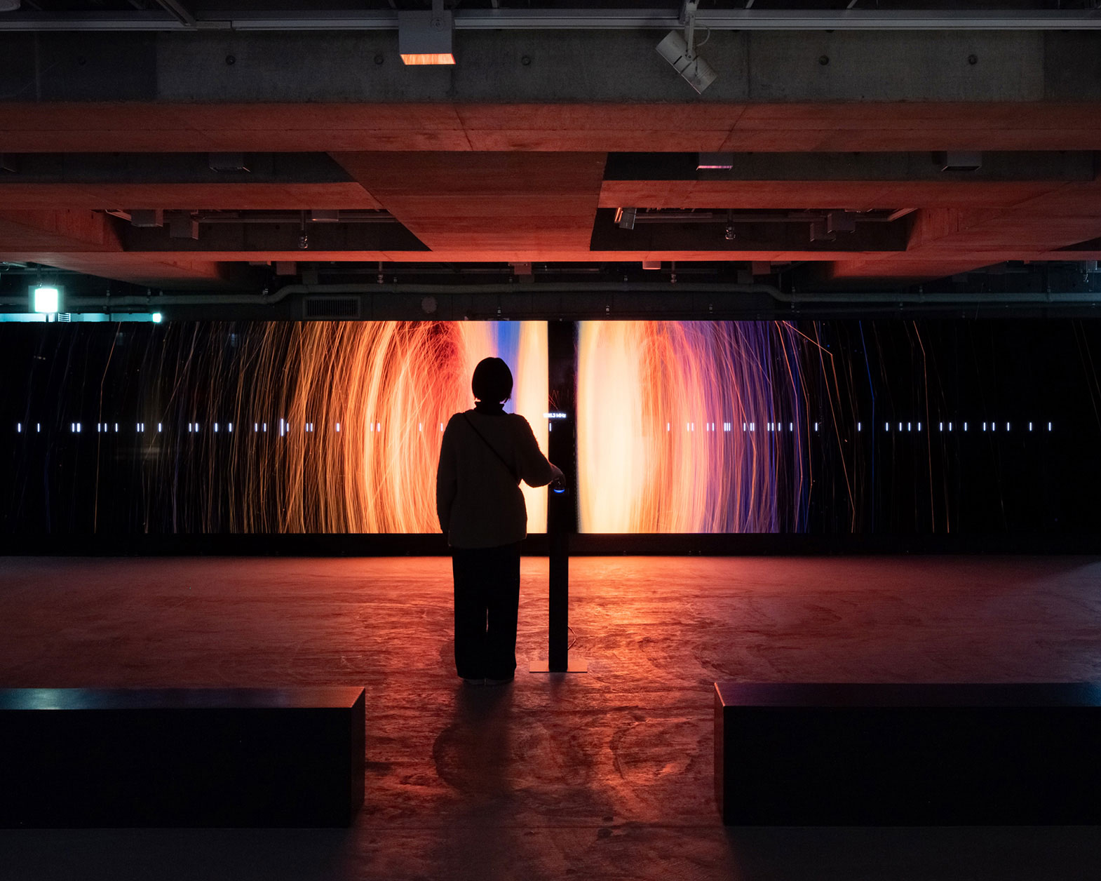 薄暗い空間の奥には赤やオレンジの映像が映し出された大きなディスプレイがあり、その前に人が一人立っている。