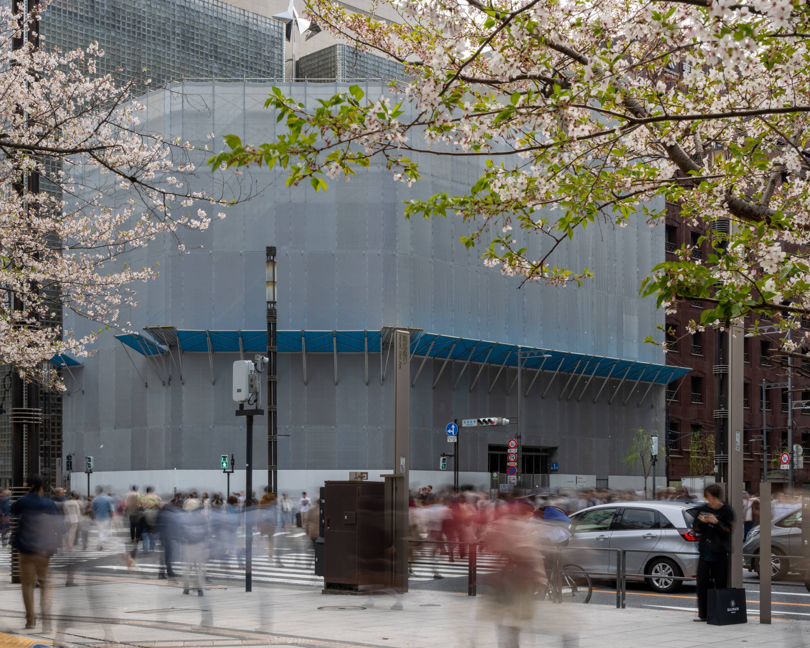 外観。塔屋の高さまで工事仮囲いに覆われたGinza Sony Parkと葉桜との間の数寄屋橋交差点は多くの人々が行き交っている。