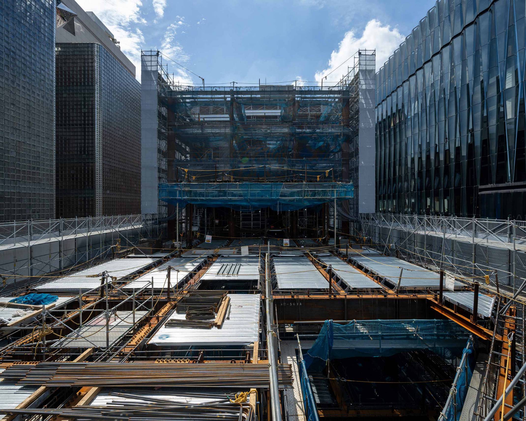 外観。コンクリート打設前の足場に囲われた屋上階の躯体工事。床面には鉄骨とデッキが露わになっている。