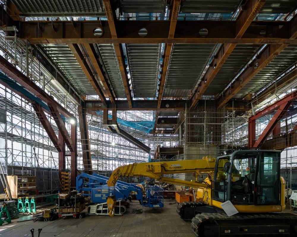 内観。仮囲い足場で囲われた地上吹き抜け部。天井には鉄骨が組まれ、地上には建築用重機が置かれている。