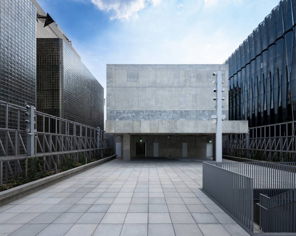 外観。周囲の建物よりも低く構えた屋上階で、青空のもとコンクリートの塔屋と東屋がある。