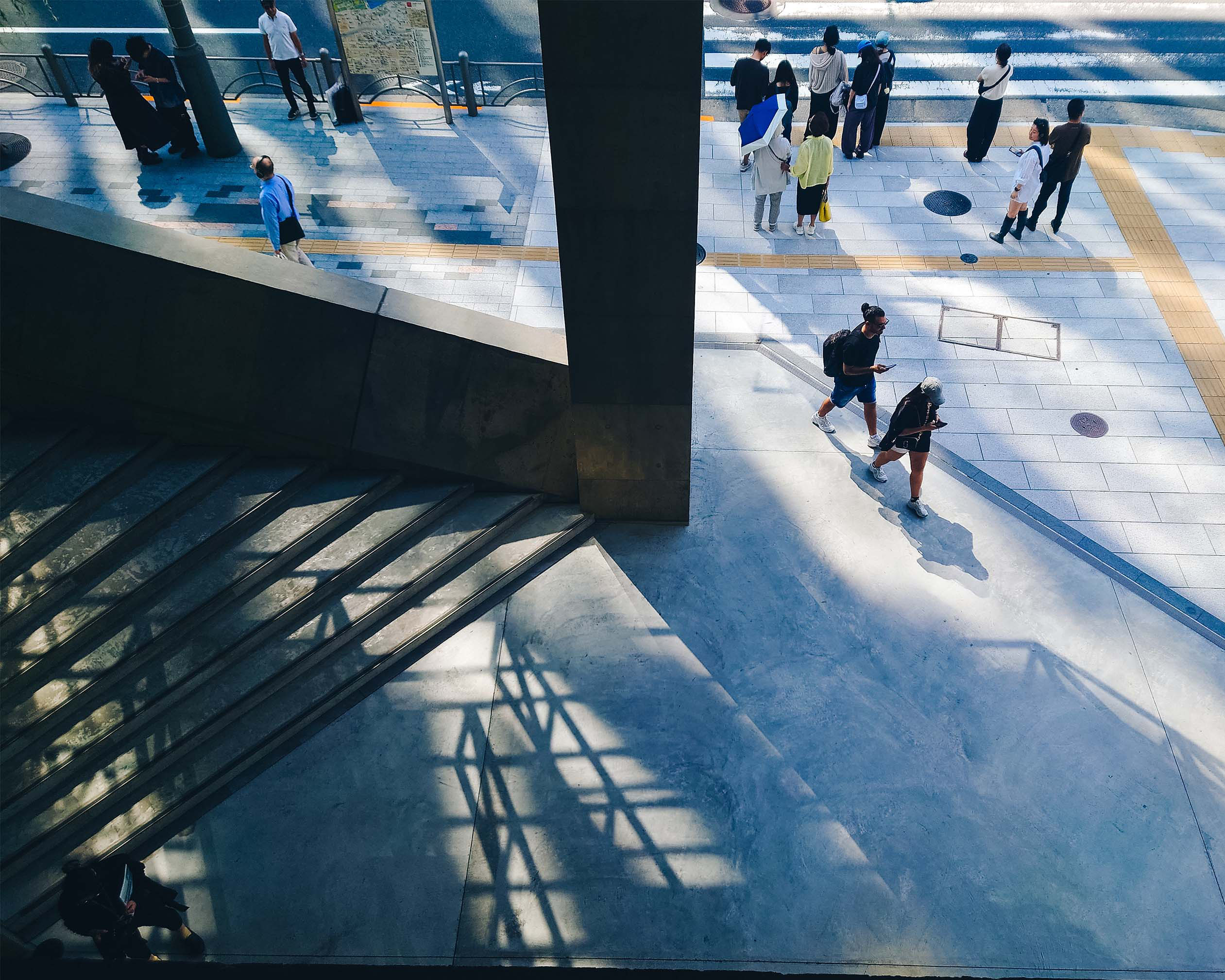Ginza Sony Parkの吹き抜け空間を上階から捉えた風景。コンクリートの階段と床に伸びる影の中、人々が行き交う様子。