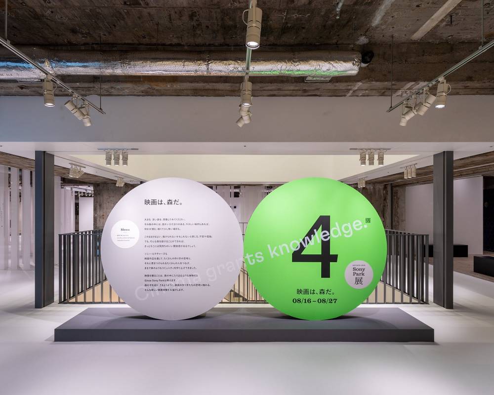 展示の様子。ステートメントの描かれたグレー色の丸いオブジェと「4」と描かれたグリーン色の丸いオブジェが地下2階の会場入口に並んでいる。