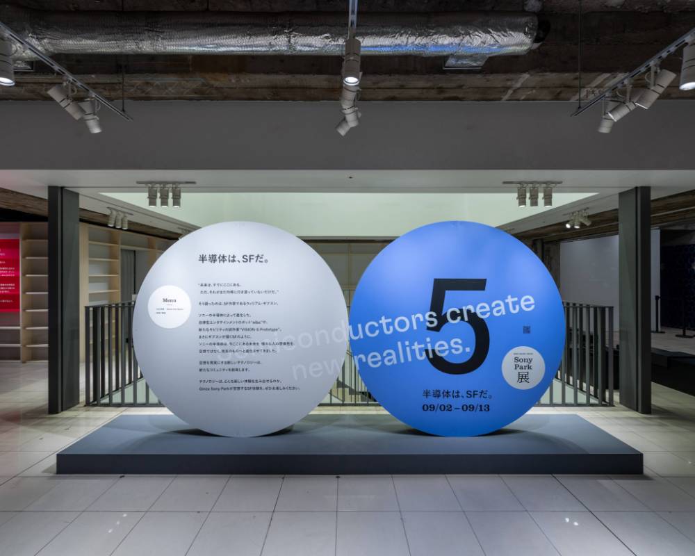 展示の様子。ステートメントの描かれたグレー色の丸いオブジェと「5」と描かれたブルー色の丸いオブジェが地下2階の会場入口に並んでいる。