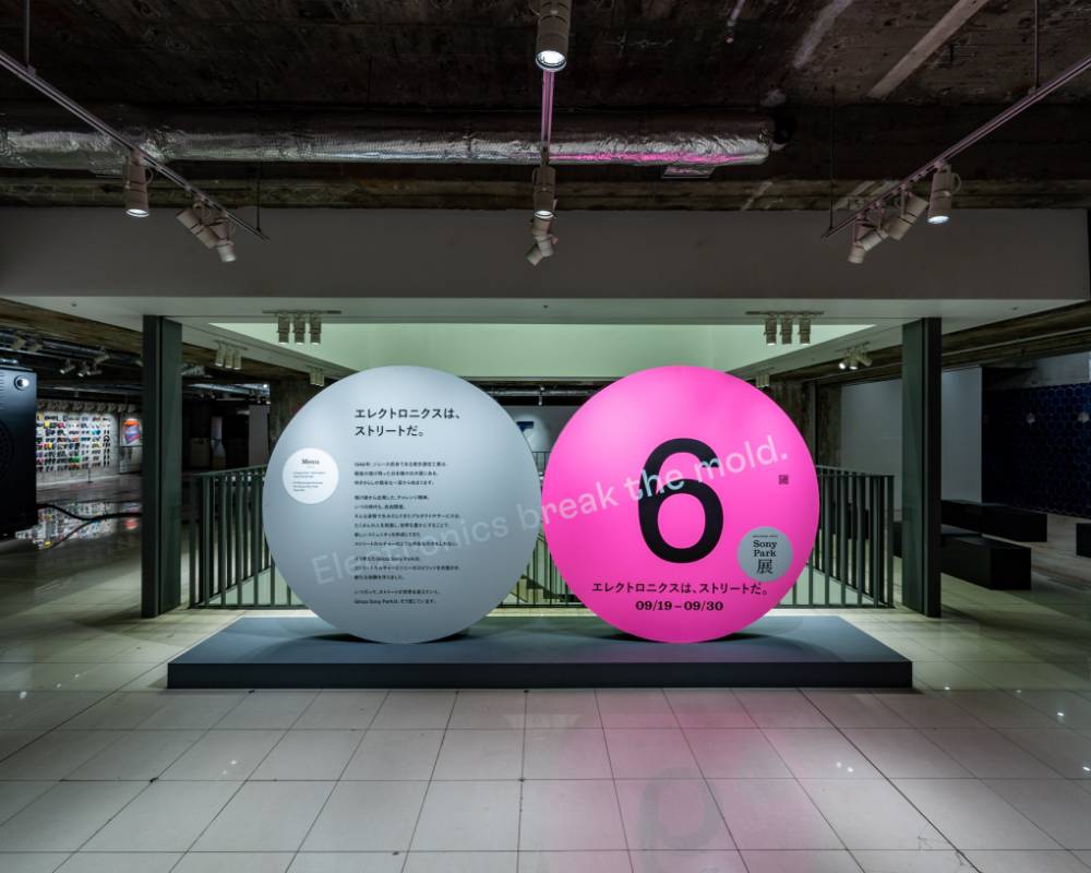 展示の様子。ステートメントの描かれたグレー色の丸いオブジェと「6」と描かれたピンク色の丸いオブジェが地下2階の会場入口に並んでいる。