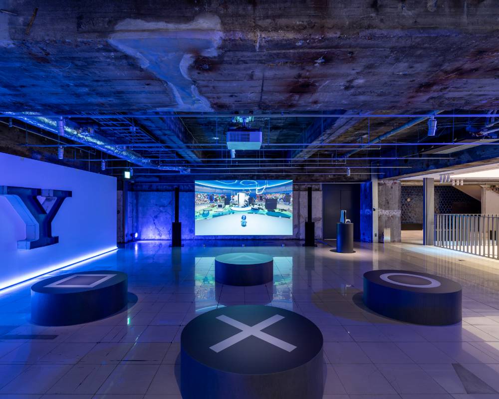 展示の様子。照明で会場が全体的にブルーになった空間で、奥の壁面にはゲーム画面が大きく投影され、手前には四角・三角・丸・バツのマークが描かれたベンチが置いてある。