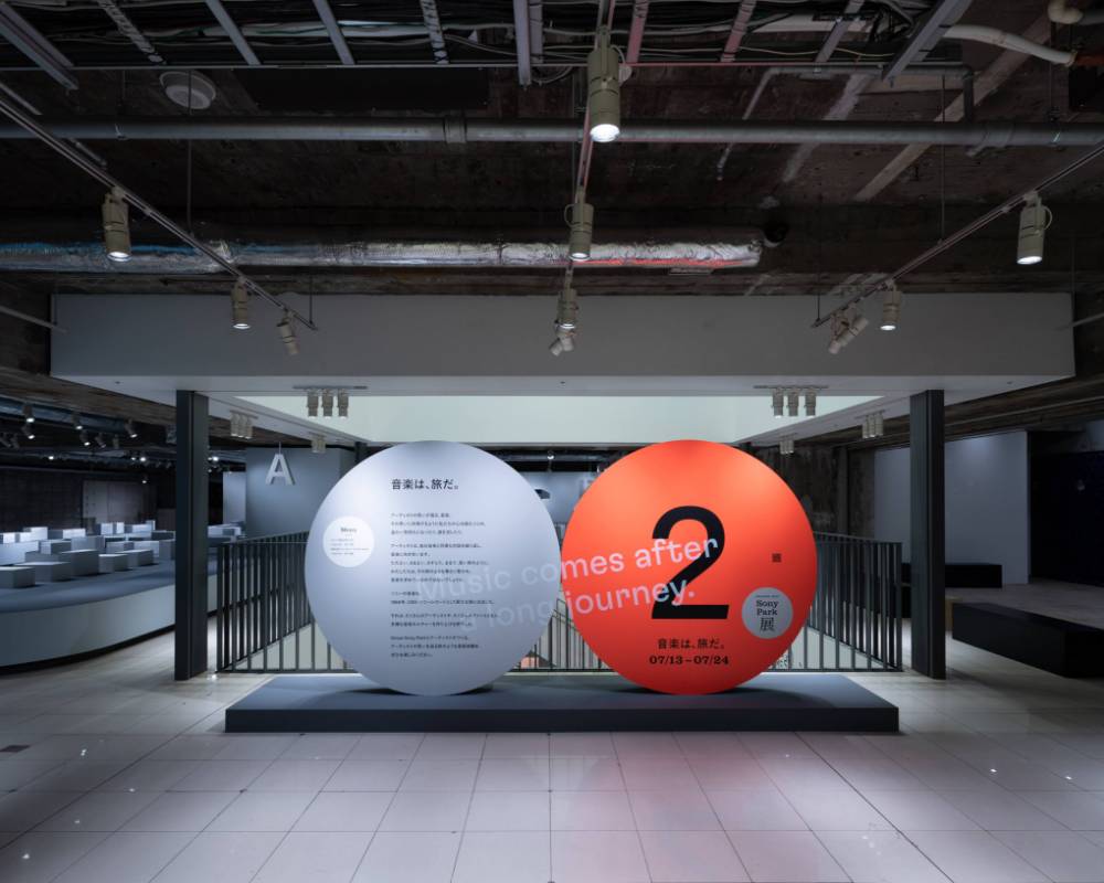 展示の様子。ステートメントの描かれたグレー色の丸いオブジェと「2」と描かれたオレンジ色の丸いオブジェが地下2階の会場入口に並んでいる。