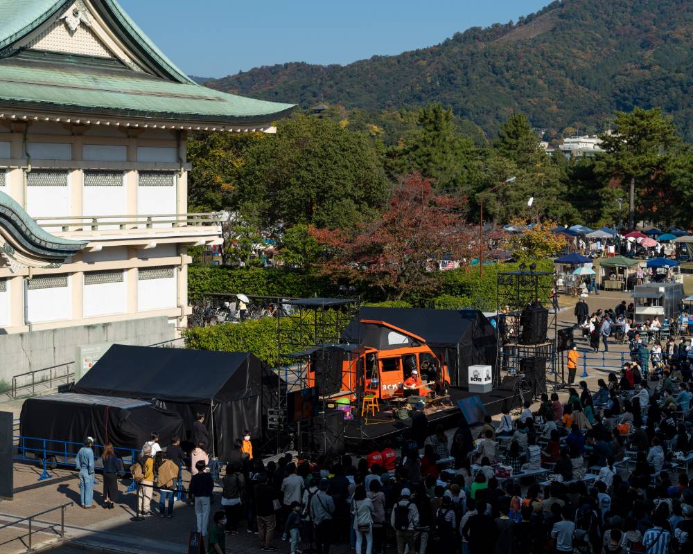 ライブパフォーマンスの様子。ロームシアター京都の屋外「ローム・スクエア」に停まったオレンジのキャンピングカーの前で演奏する奥田民生のライブパフォーマンスに多くの人々が集まる。