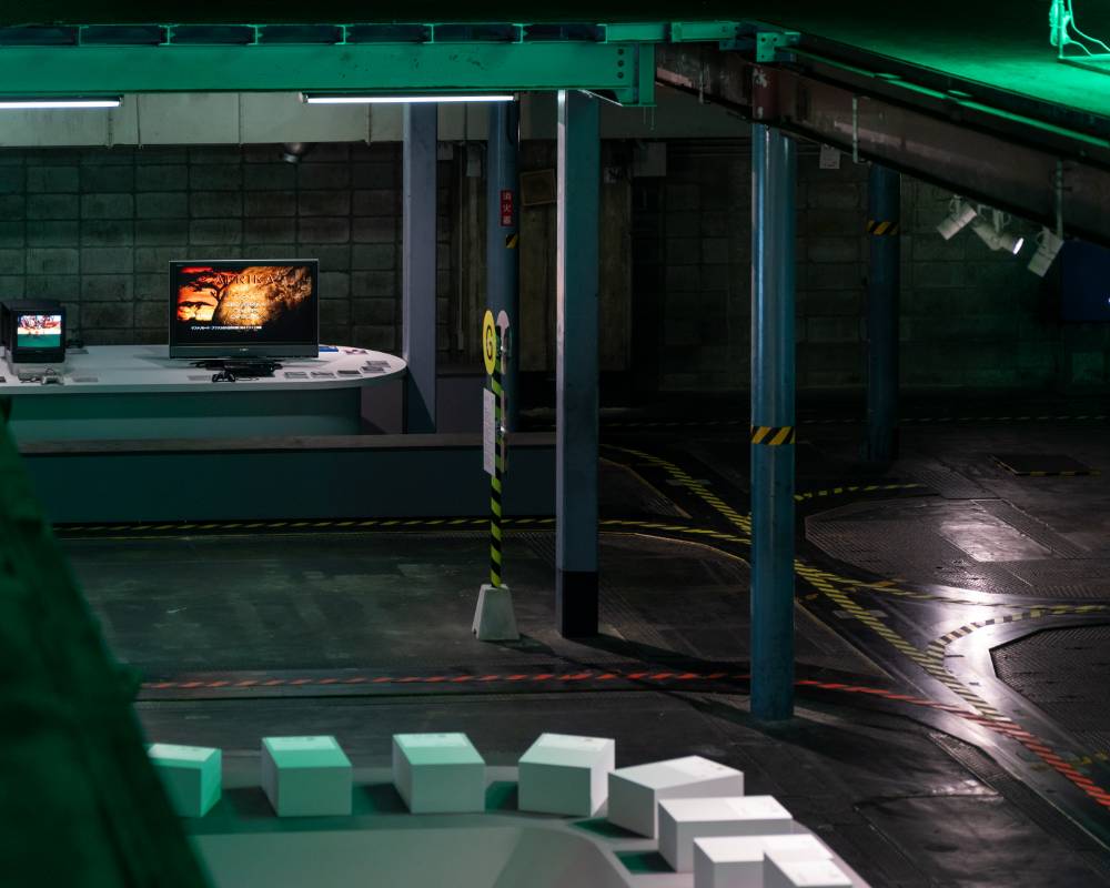 展示の様子。会場を俯瞰した奥にはゲームタイトルの映ったモニターが、手前には複数の白いBOXが展示されている。