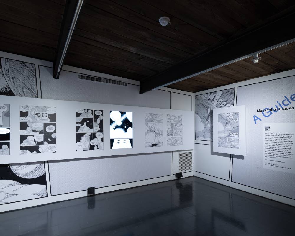 展示の様子。壁面の大きなグラフィックの上にマンガ『案内人』が１ページずつ展示され、ところどころモニターが埋め込まれたコマで映像が映し出されている。