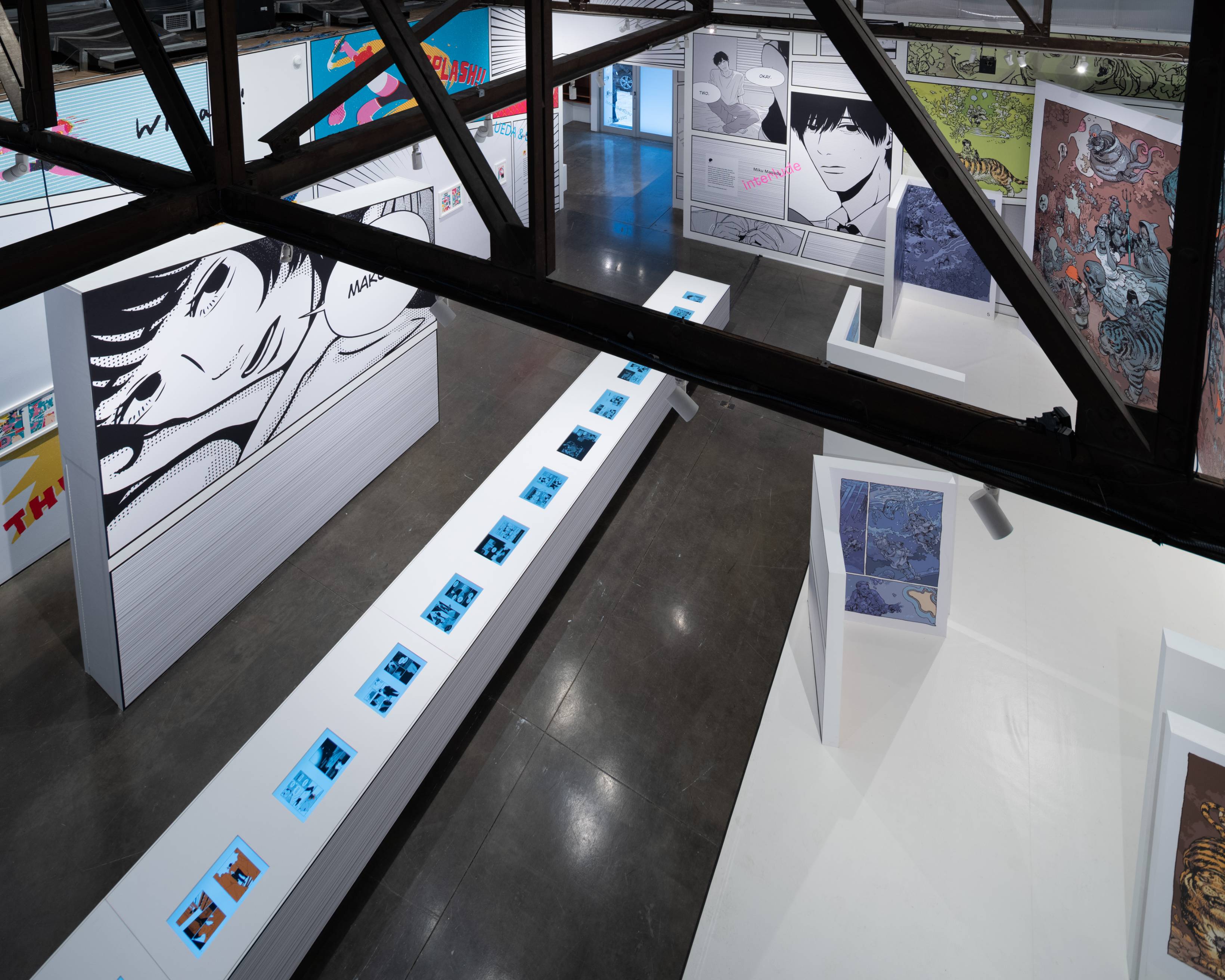 展示の様子。会場1階を俯瞰すると大きくマンガが展示された壁やパーティション、そして複数のモニターが埋め込まれた長いテーブルがある。