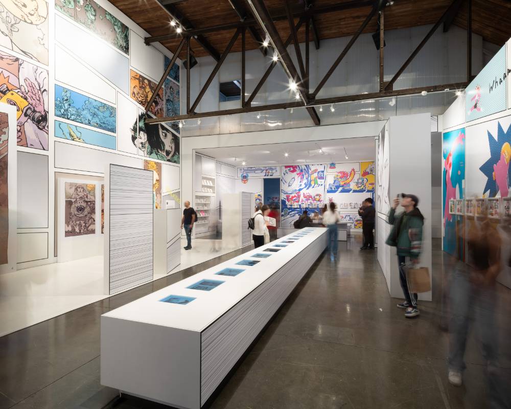 内観。いくつものモニターが埋め込まれた長いテーブルが伸びる会場中央のまわりで来場者が賑わっている。