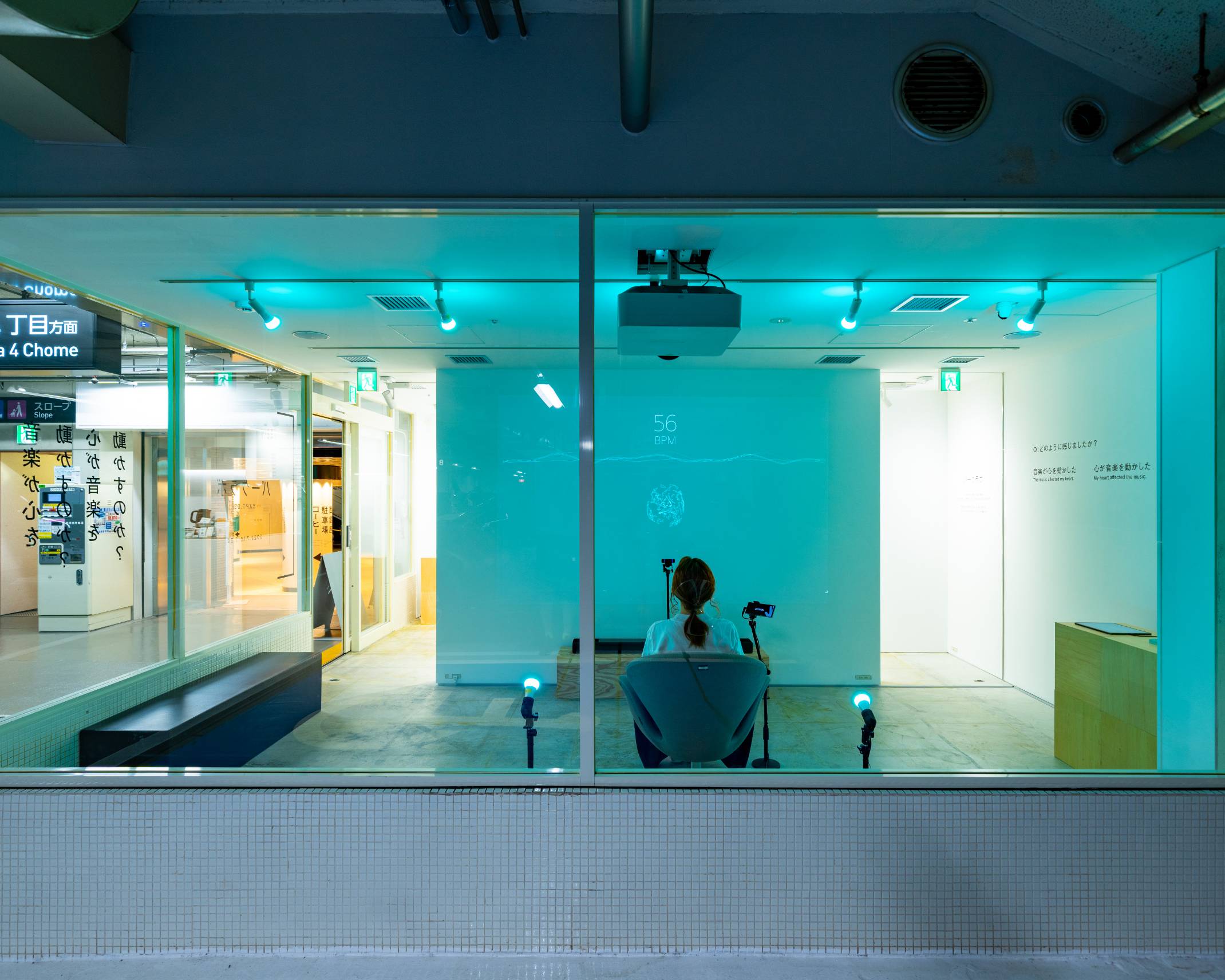 体験の様子。来場者は椅子に座り、正面の壁には映像が投影されている。空間全体は照明によってブルーになっている。