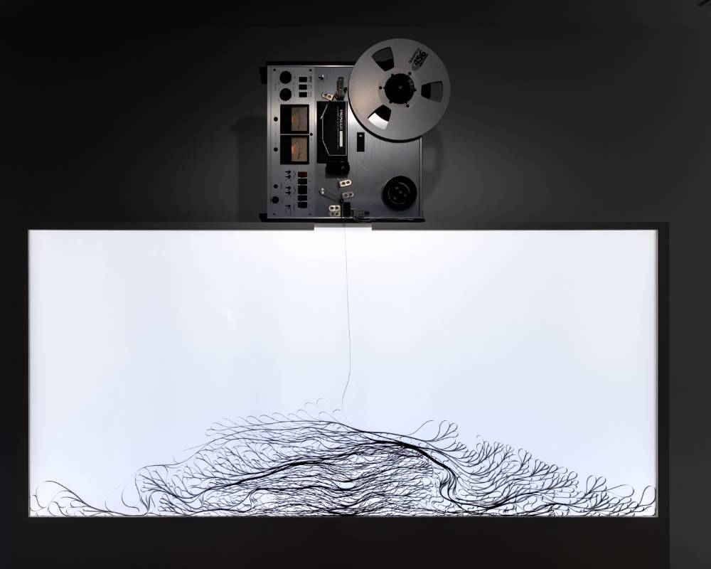 展示の様子。オープンリール式のテープレコーダーから落ちるテープが、折り重なって容器内に溜まっている。