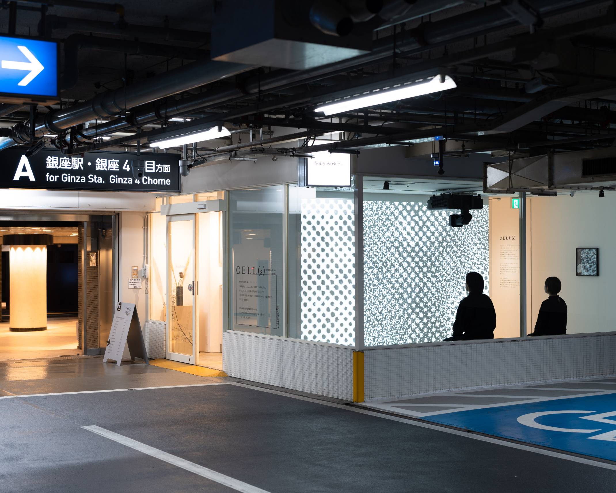 外観写真。展示スペース内の壁面に投影された作品を来場者２名が座って眺めている。