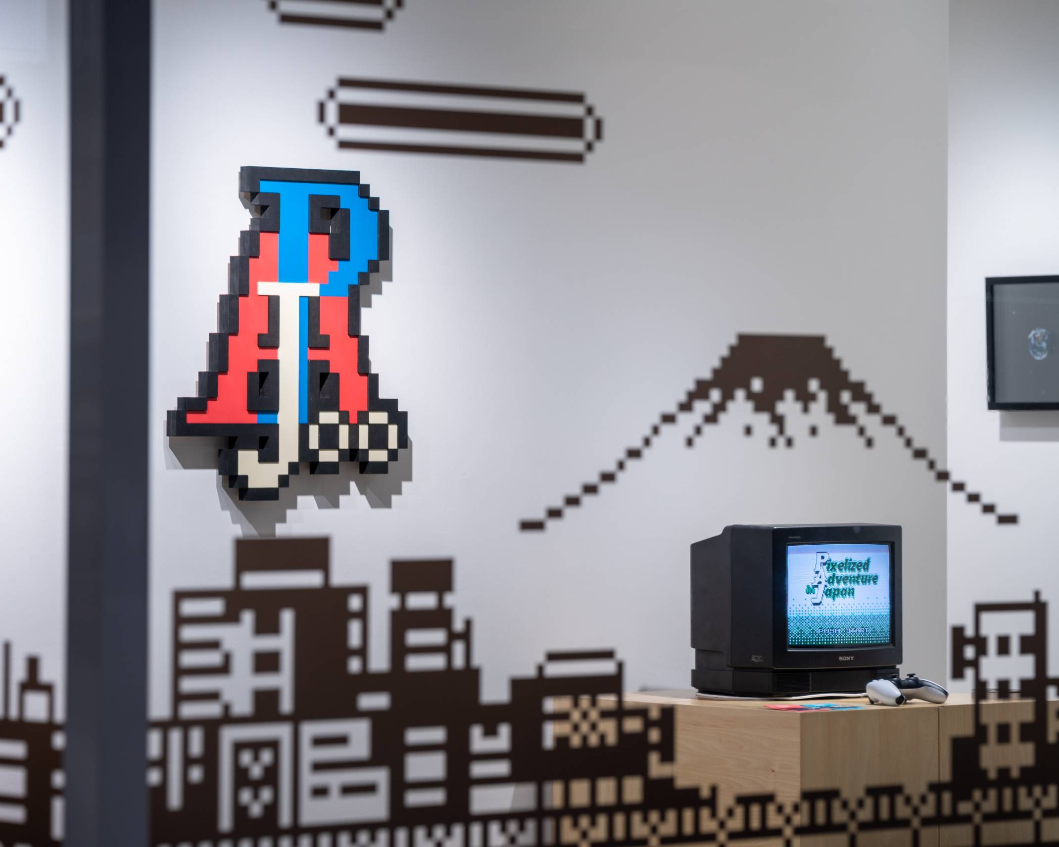 展示の様子。壁面にはピクセルアートが飾られ、台の上にはゲーム画面が映し出されたブラウン管テレビとコントローラーが置かれている。
