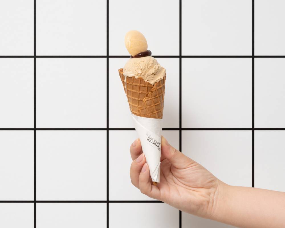 商品。格子柄の壁の前で右手に持った、コーンに盛り付けられたモカ色のアイスクリーム。