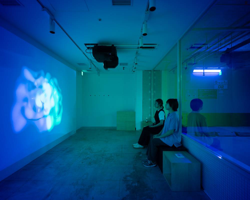 体験の様子。頭部にデバイスをつけた２名の来場者が着席し、壁面に投影された映像を見ている。