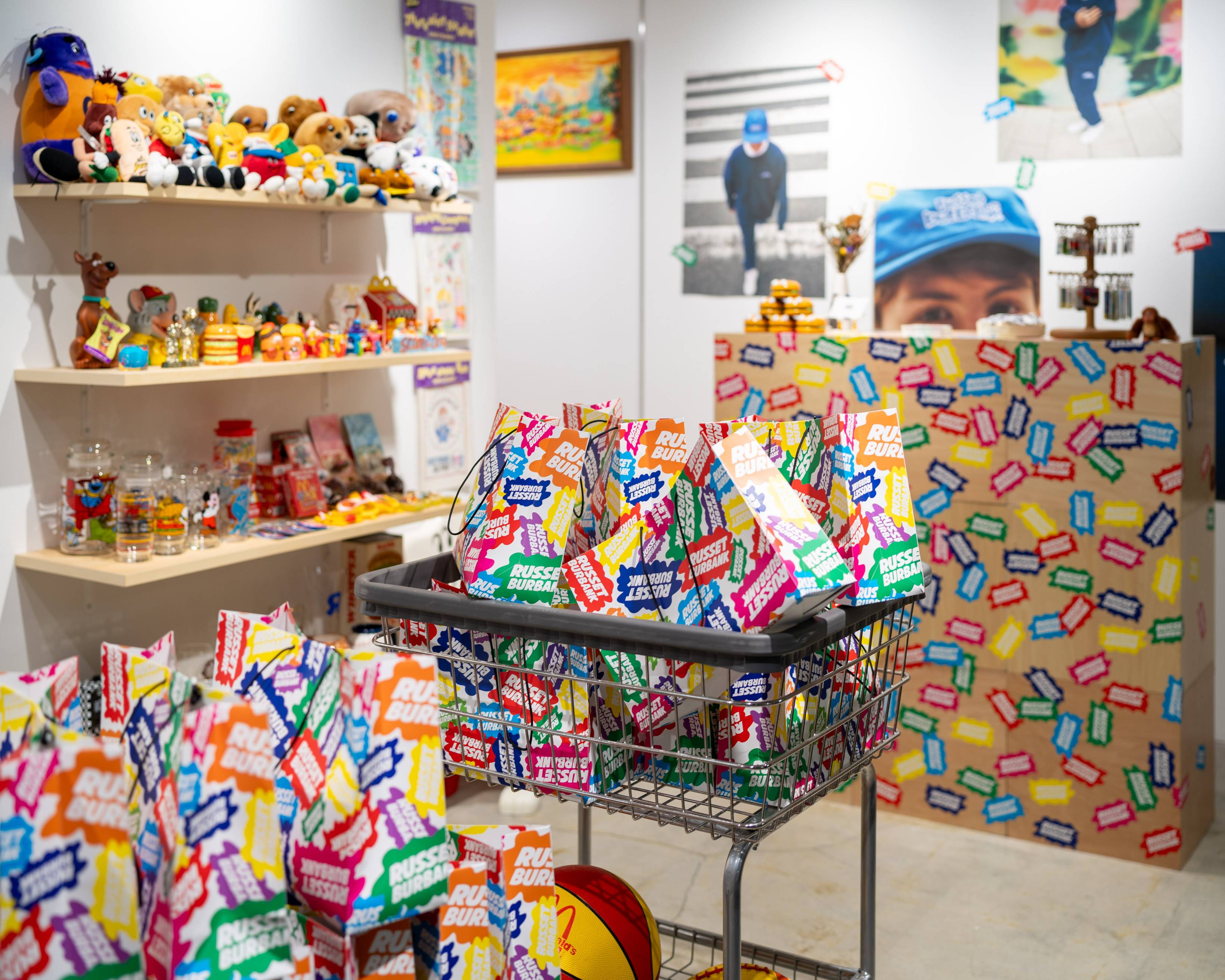 展示の様子。壁面の棚にはさまざまなヴィンテージトイが並び、中央のカートにはカラフルな紙袋が詰められている。