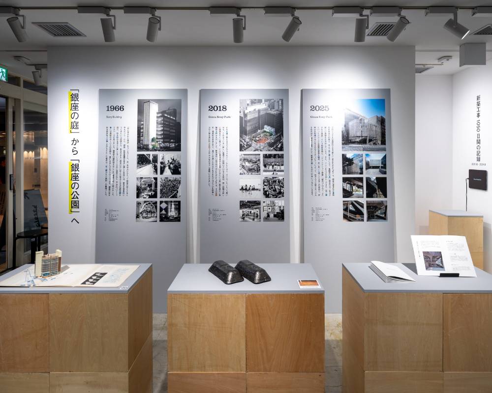 展示の様子。壁面には縦長のパネルが3枚並び、手前の3つの台の上には冊子や金属の塊が展示されている。