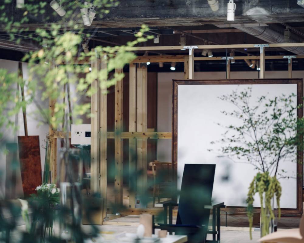 展示の様子。枝物の植栽越しに、大きな白い絵画や黒い椅子などの複数の椅子が展示されている。