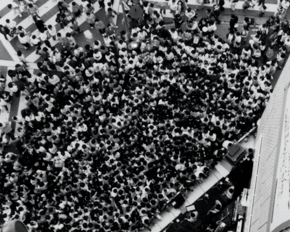 ライブパフォーマンスの様子（モノトーン）。ソニースクエア（屋外イベントスペース）の音楽ライブに集まる人々で歩道が埋め尽くされている。