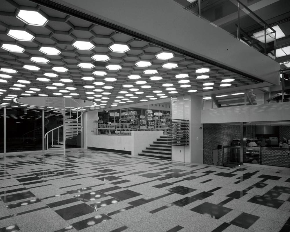 内観（モノトーン）。開業当時の1階エントランスホール。左奥には螺旋階段があり、天井にはハニカム構造の照明が広がっている。