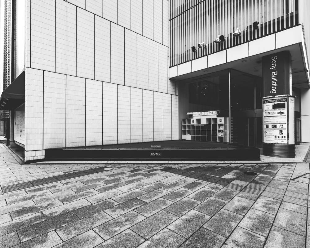 外観（モノトーン）。手前が歩道に隣接して開かれている三角形の広場。歩道より一段高くなった屋外イベントスペースになっている。