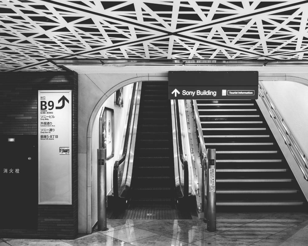 地下外観（モノトーン）。ロマネスク様式の厚い壁と半円アーチ型天井に囲まれた空間にエスカレーターと階段があり、入口には地下鉄のサインボードがある。