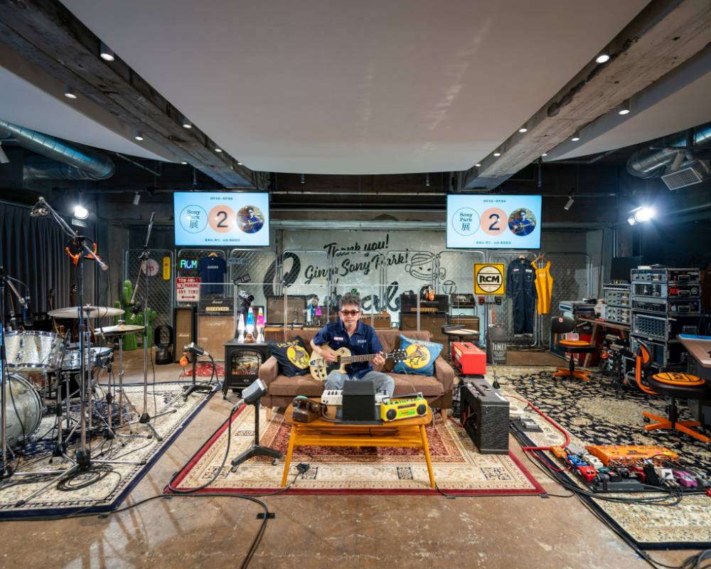 展示／ライブパフォーマンスの様子。宅録スタイルのレコーディングスペースをステージ上に再現した空間で、中央に設置されたソファーに奥田民生がギターを持って座っている。