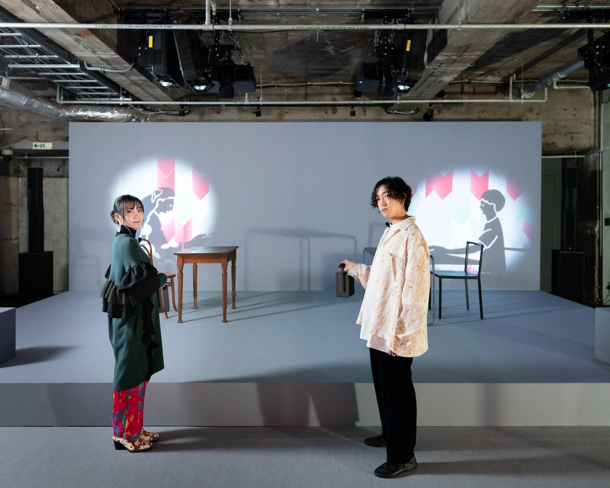 体験の様子。特別な電灯で壁面を照らすと影絵のアニメーションが浮かび上がる展示をYOASOBIの2人が体験している。