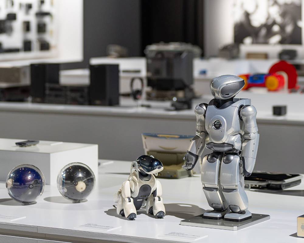 展示の様子。手前の展示台に、エンタテインメントロボット「aibo」、小型二足歩行エンターテインメントロボット「QRIO」や球体型の「Q.taro」が展示されている。