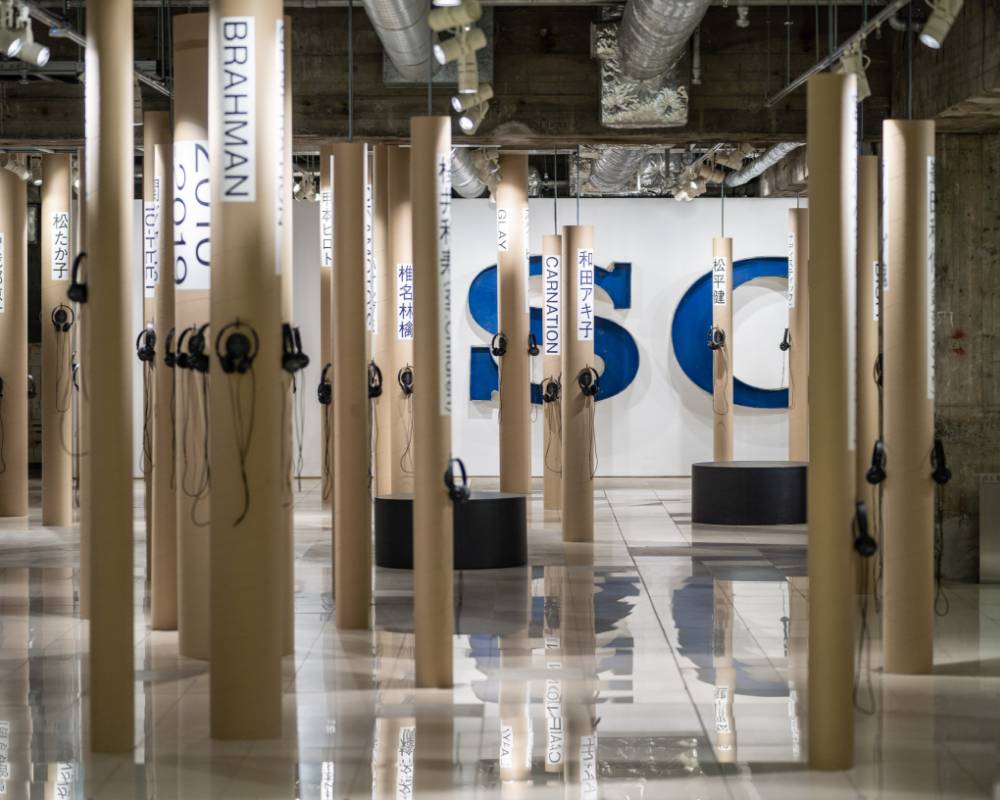 展示の様子。アーティスト名の書かれた複数の紙管が地下空間に森の木々のように立てられ、それぞれヘッドフォンが掛けられている。