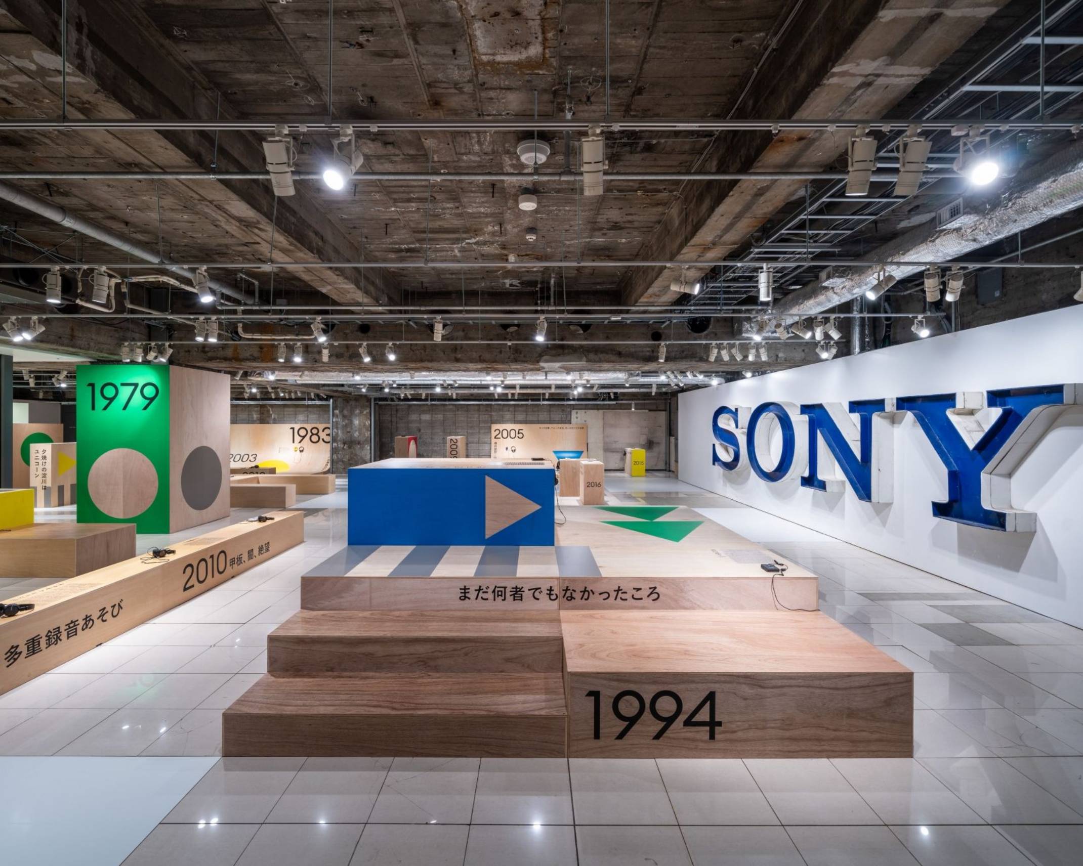 展示の様子。右手壁面に「SONY」の大きなサインがある地下会場には、年号の描かれた公園の遊具のように組まれた木製の展示台があり、それぞれの年代のウォークマンが展示されている。
