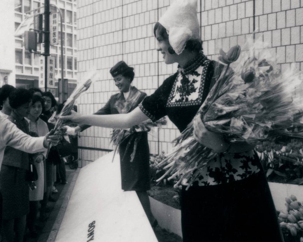 イベントの様子（モノトーン）。ソニースクエア（屋外イベントスペース）でオランダの民族衣装を着た女性と客室乗務員がチューリップを配布している。